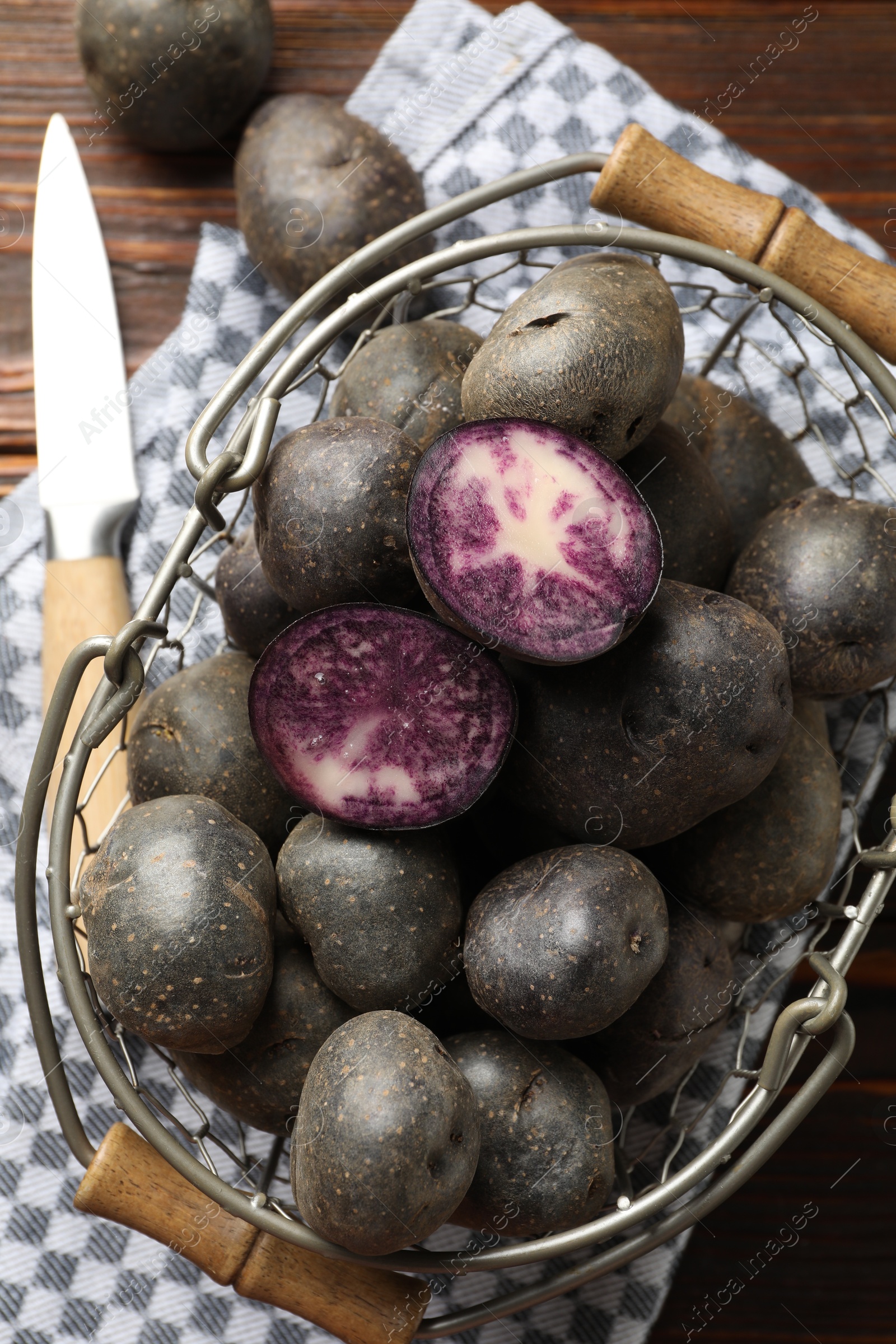 Photo of Raw purple potatoes in metal basket and knife on wooden table, flat lay