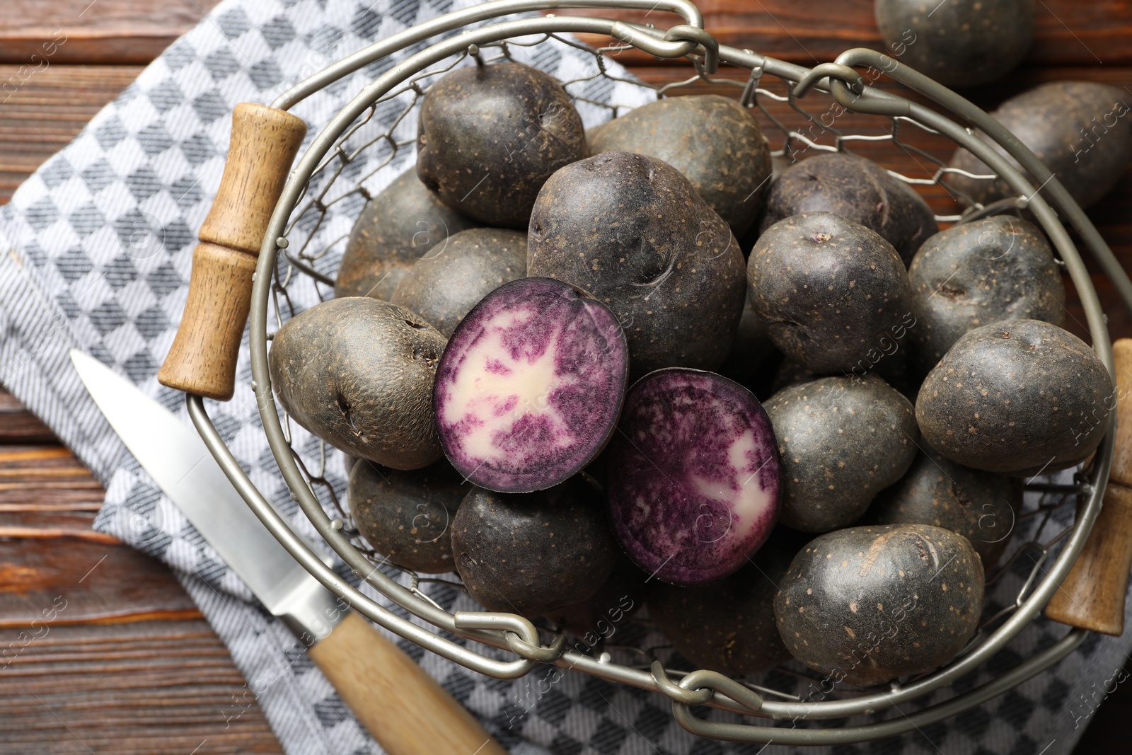 Photo of Raw purple potatoes in metal basket and knife on wooden table, flat lay