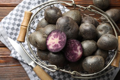 Photo of Raw purple potatoes in metal basket and knife on wooden table, flat lay