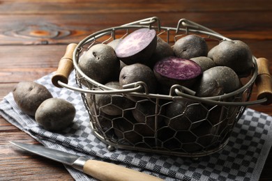 Photo of Raw purple potatoes in metal basket and knife on wooden table, closeup