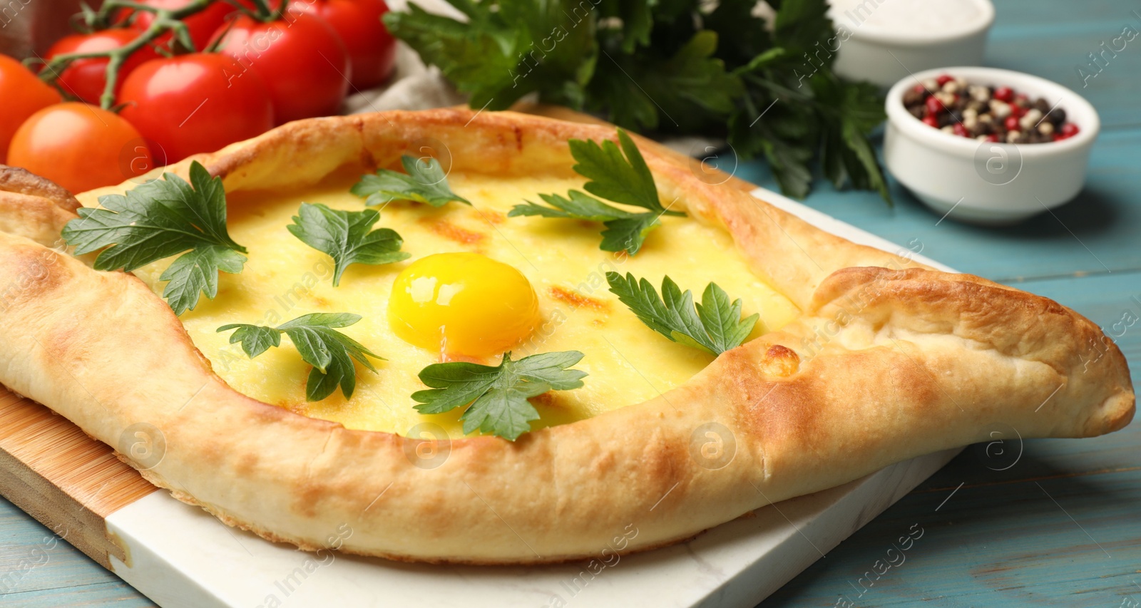 Photo of Tasty khachapuri with cheese, egg, parsley, tomatoes and spices on light blue wooden table, closeup