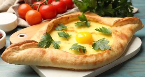 Photo of Tasty khachapuri with cheese, egg, parsley, tomatoes and spices on light blue wooden table, closeup
