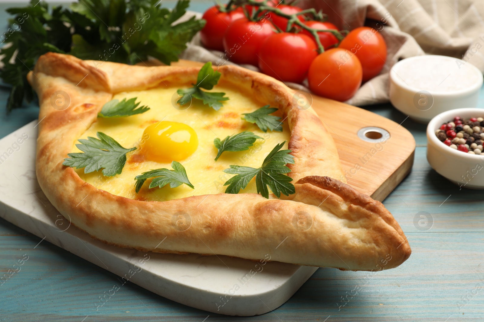 Photo of Tasty khachapuri with cheese, egg, parsley, tomatoes and spices on light blue wooden table, closeup