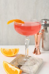 Photo of Martini cocktail with garnish, slices of orange and bartender equipment on white table, closeup