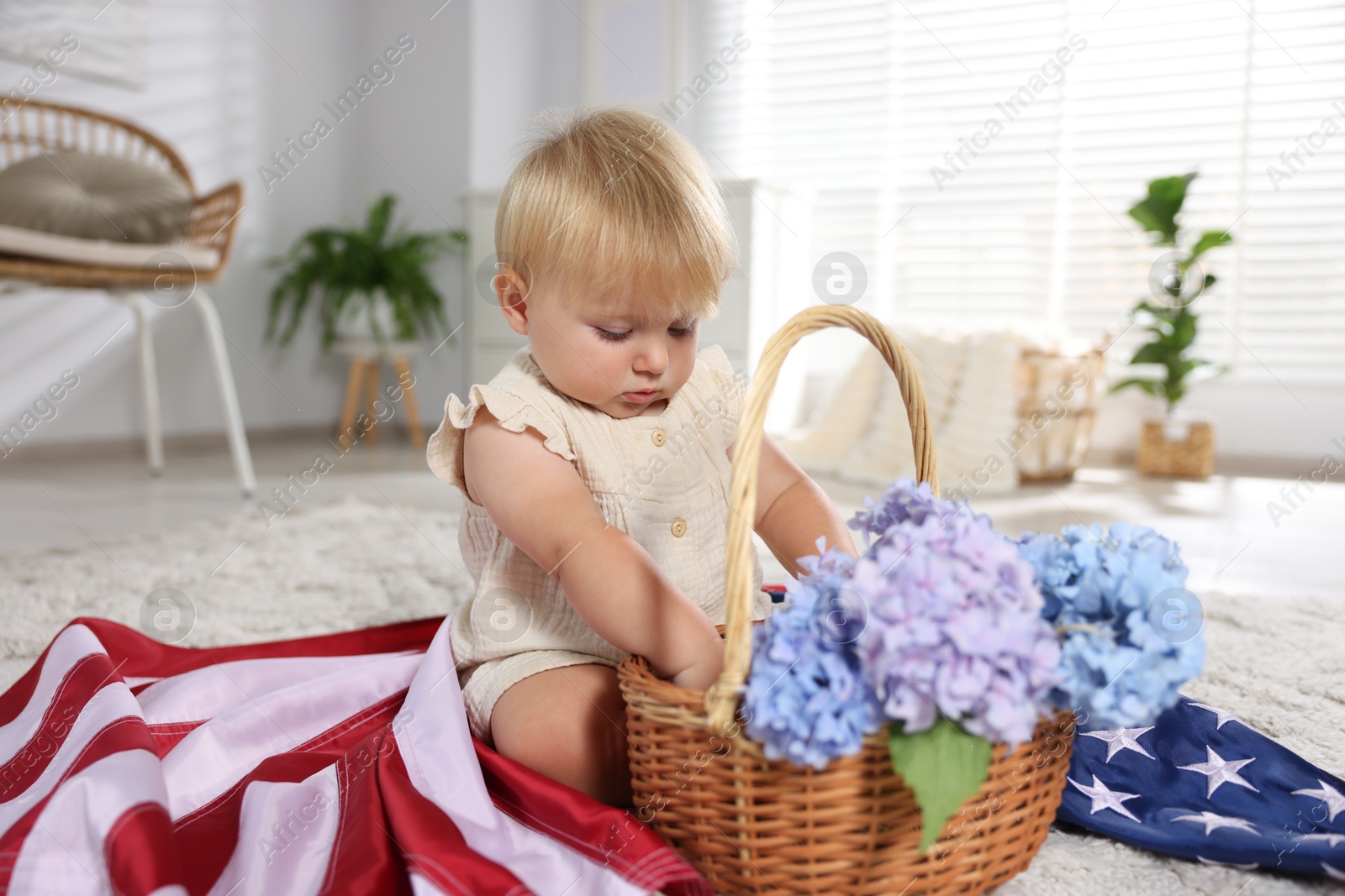 Photo of Cute little child with flag of USA and wicker basket of hydrangea flowers on floor at home