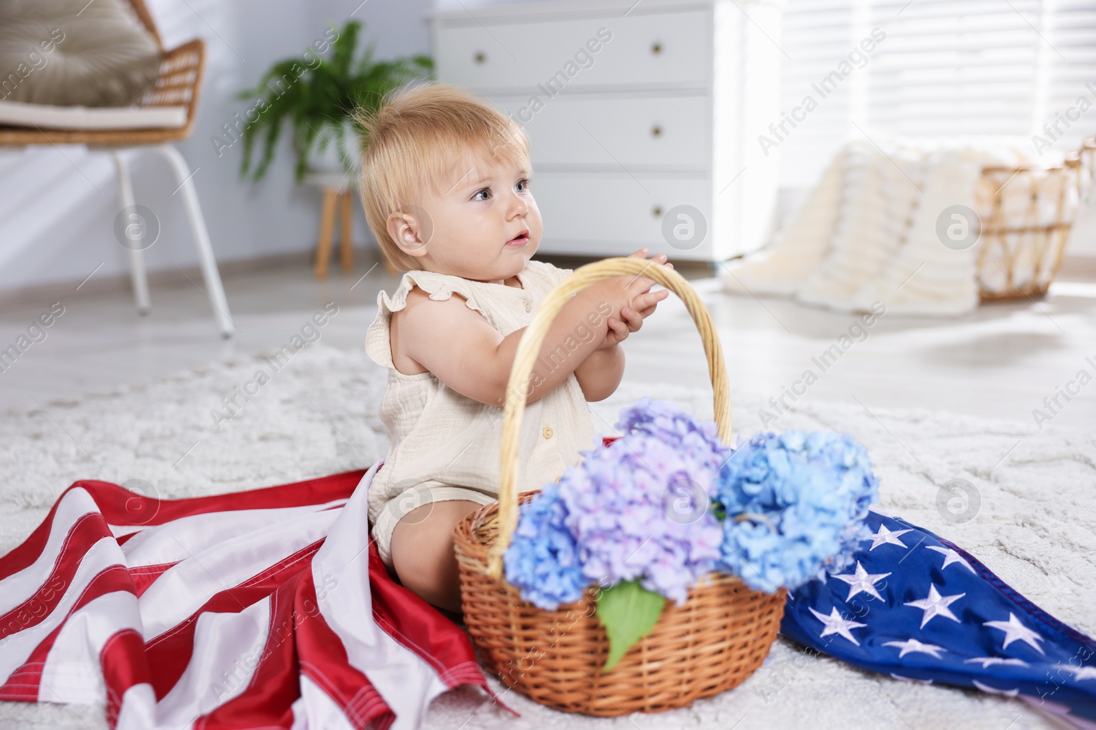 Photo of Cute little child with flag of USA and wicker basket of hydrangea flowers on floor at home