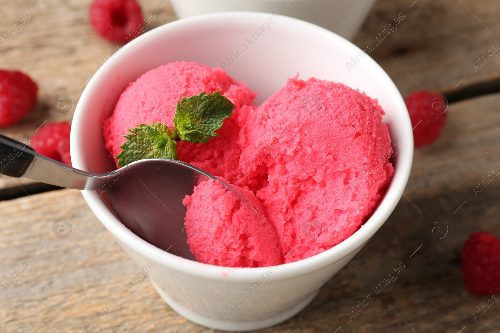 Photo of Taking delicious raspberry sorbet with mint from bowl with spoon at wooden table, closeup