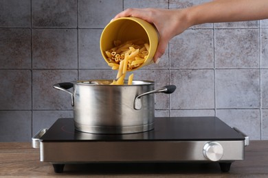 Photo of Woman putting penne pasta into pot with boiling water on electric stove, closeup