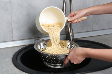 Photo of Woman draining boiled tagliatelle pasta into colander at sink in kitchen, closeup