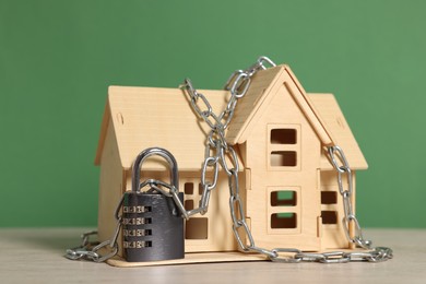 Photo of Home security. House model with chain and padlock on wooden table against green background, closeup