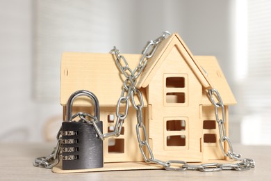 Photo of Home security. House model with chain and padlock on wooden table indoors, closeup