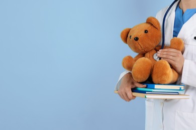 Photo of Pediatrician with notebooks checking heartbeat of toy bear on light blue background, closeup. Space for text
