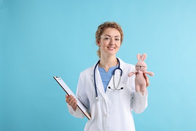 Photo of Professional pediatrician with toy bunny and clipboard on light blue background