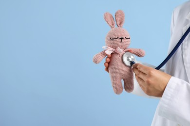 Photo of Professional pediatrician checking heartbeat of toy bunny on light blue background, closeup. Space for text