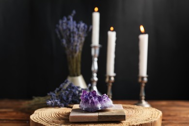 Photo of Amethyst gemstone on wooden table against lavender and burning candles, selective focus