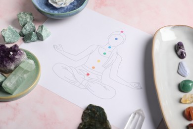 Photo of Drawing of chakras on human body and gemstones on light pink table, closeup
