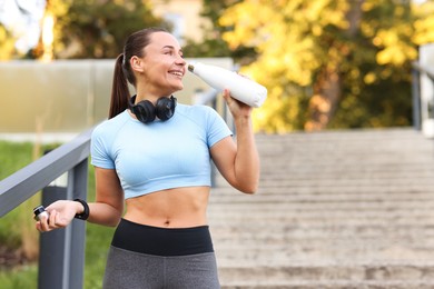 Photo of Athletic young woman with headphones drinking water before running outdoors, space for text