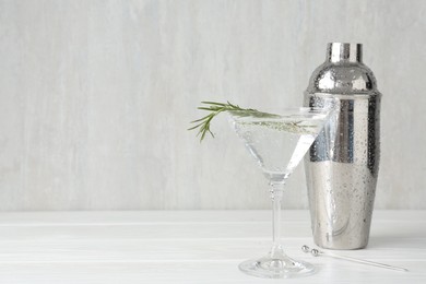 Photo of Martini cocktail with rosemary in glass and shaker on white wooden table, closeup. Space for text