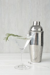 Photo of Martini cocktail with rosemary in glass and shaker on white wooden table, closeup