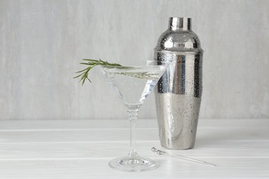 Photo of Martini cocktail with rosemary in glass and shaker on white wooden table, closeup