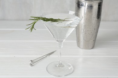 Photo of Martini cocktail with rosemary in glass and shaker on white wooden table, closeup