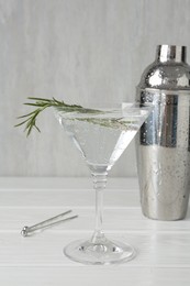 Photo of Martini cocktail with rosemary in glass and shaker on white wooden table, closeup
