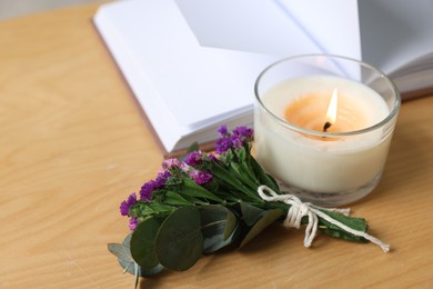 Photo of Burning candle, open book and flowers on wooden table indoors, closeup. Space for text