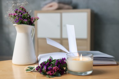 Photo of Burning candle, open book and flowers on wooden table indoors, closeup