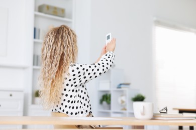 Photo of Woman using remote to control air conditioner in office, back view