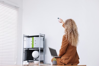 Photo of Woman using remote to control air conditioner in office, back view