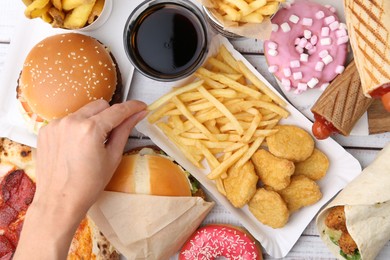 Photo of Woman taking tasty French fry at light wooden table with different fast food and cola, top view