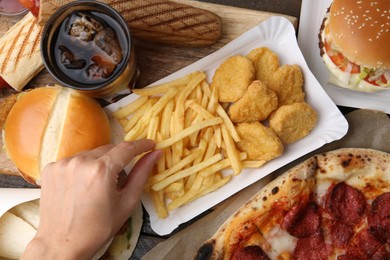 Photo of Woman taking tasty French fry at wooden table with different fast food and cola, top view