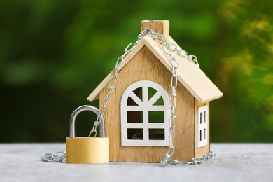 Photo of Home security. House model, chain and padlock on light grey table outdoors, closeup