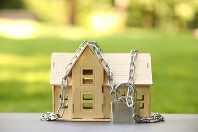 Photo of Home security. House model, chain and padlock on light table outdoors, closeup