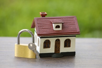 Photo of Home security. House model, key and padlock on wooden table outdoors, closeup
