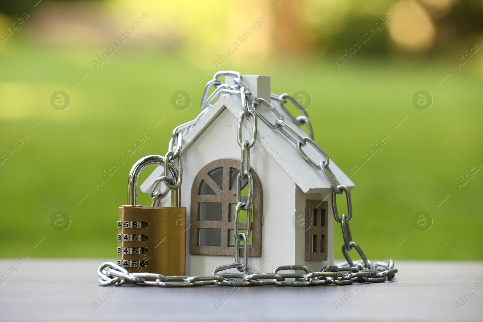 Photo of Home security. House model, chain and padlock on light grey table outdoors, closeup