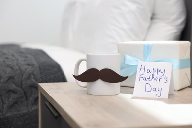 Photo of Happy Father's Day card, cup with mustache and gift on wooden table at home, closeup. Space for text