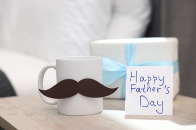 Photo of Happy Father's Day card, cup with mustache and gift on wooden table at home, closeup