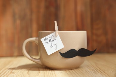 Photo of Happy Father's Day. Cup with mustache and note on wooden table, closeup