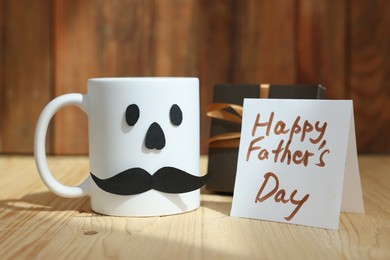 Photo of Happy Father's Day card, cup with paper face and gift on wooden table, closeup