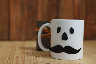 Photo of Happy Father's Day. Cup with paper face and gift on wooden table, closeup. Space for text