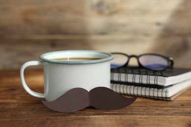 Photo of Happy Father's Day. Cup with mustache, notebooks and glasses on wooden table, closeup