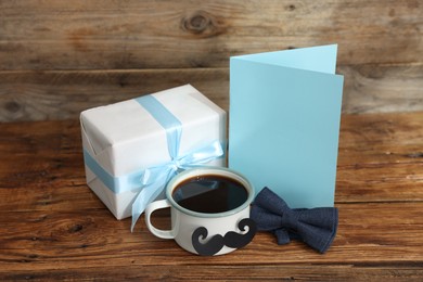 Photo of Happy Father's Day. Cup with mustache, card and gift on wooden table. Space for text