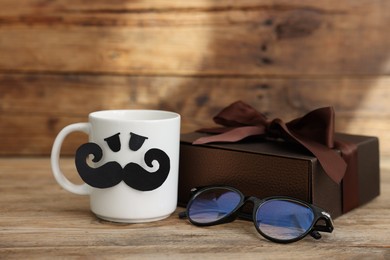 Photo of Happy Father's Day. Cup with paper face, gift and glasses on wooden table