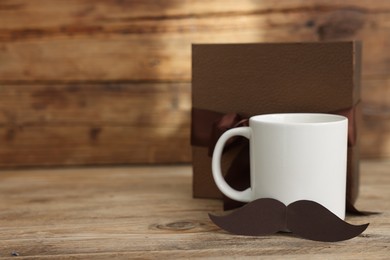 Photo of Happy Father's Day. Cup with mustache and gift on wooden table. Space for text