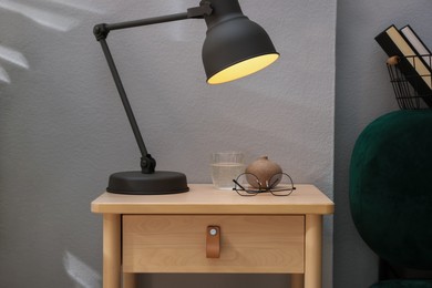 Photo of Wooden side table with glass of water, lamp and glasses indoors, closeup