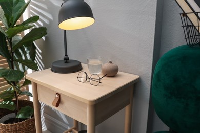Photo of Wooden side table with glass of water, lamp and glasses indoors