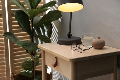 Photo of Wooden side table with glass of water, lamp and glasses indoors, closeup