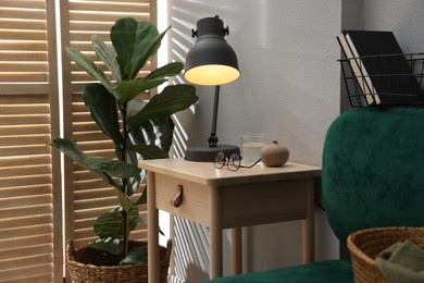 Photo of Wooden side table with glass of water, lamp and glasses indoors