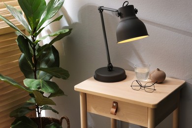 Photo of Wooden side table with glass of water, lamp and glasses indoors, closeup